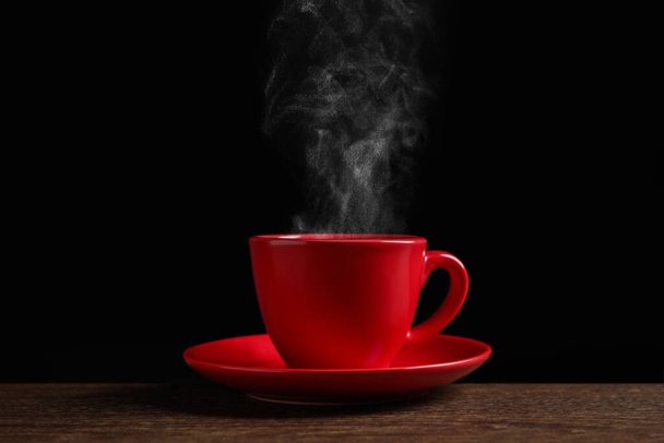 Red cup with hot steaming coffee on wooden table against black background - Valokuva, kuva