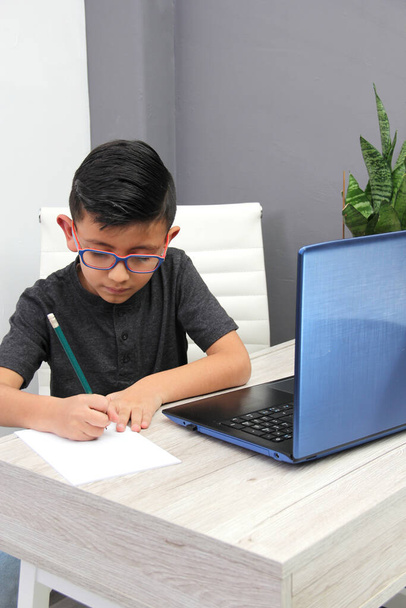 7-year-old Latino αγόρι με γυαλιά κάνει κατ 'οίκον εκπαίδευση παίρνει online μαθήματα στο σπίτι σε ένα γραφείο με ένα φορητό υπολογιστή, σπουδές, είναι έκπληκτος και συμμετέχει στην τάξη - Φωτογραφία, εικόνα