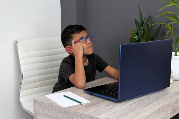 7-летний латиноамериканец в очках делает домашнее обучение берет уроки дома на рабочем столе с ноутбуком, учится, удивляется и участвует в занятиях - Фото, изображение