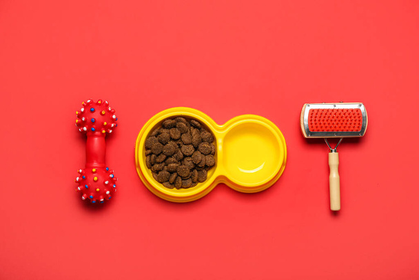 Bowl of dry pet food, grooming brush and rubber toy on red background - Foto, imagen
