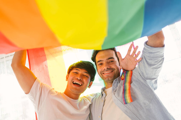 Glückliches kaukasisches und asiatisches LGBT-Paar, stehend und schwenkend unter der Regenbogenfahne LGBT Pride gemeinsam im heimischen Wohnzimmer. Vielfalt der LGBT-Beziehungen. Ein Konzept für homosexuelle Paare. - Foto, Bild