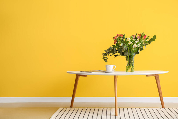 Wazon z pięknymi alstroemeria kwiaty, filiżanka kawy i czasopisma na stole w pobliżu żółtej ściany - Zdjęcie, obraz