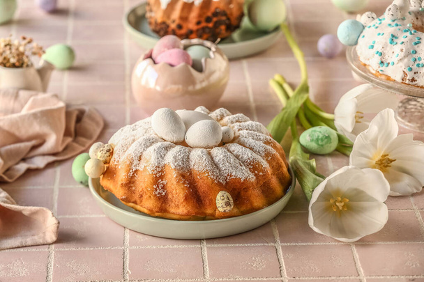 Plate with tasty Easter cake, painted eggs and beautiful tulip flowers on pink tile table - Fotografie, Obrázek