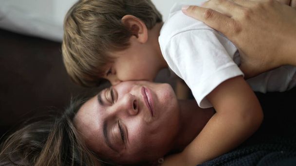 Child embracing mother lying on sofa indoors. Small boy laid on top of mom hugging. Parent and kid care affection concept - Фото, зображення