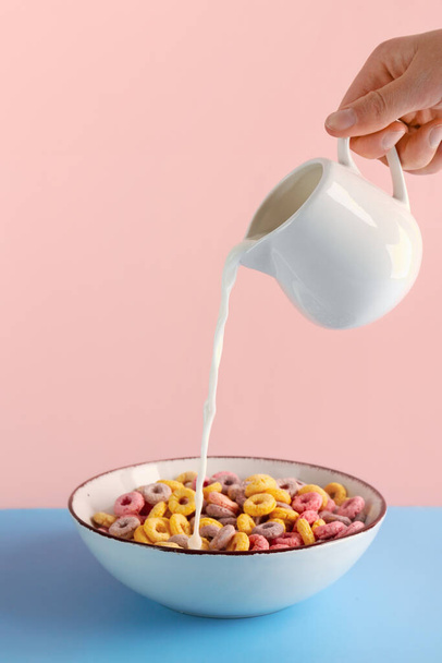 Female hand pouring milk from pitcher into bowl with colorful cereal rings on blue table against pink background - Photo, Image