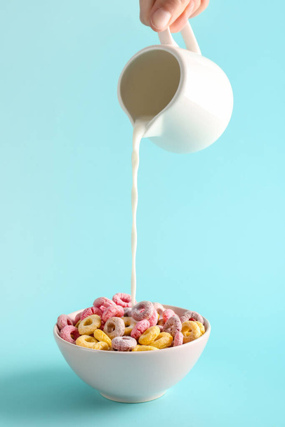 Female hand pouring milk from pitcher into bowl with colorful cereal rings on turquoise background - Photo, Image