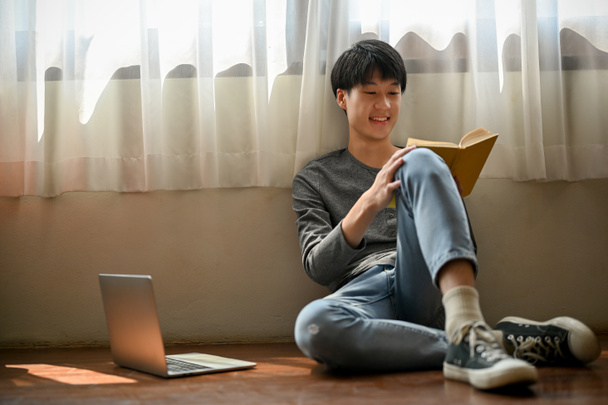 Happy and cute young Asian man in casual clothes sits on the floor in his room and enjoys reading a book. leisure or hobby concept - Zdjęcie, obraz