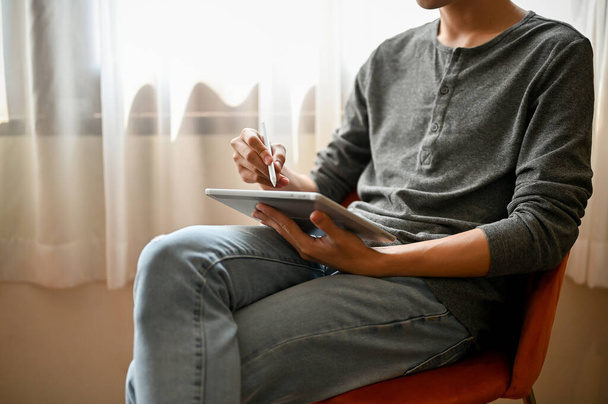 Imagem cortada de um homem asiático em roupas casuais usando seu tablet digital enquanto relaxa em uma cadeira em sua sala de estar. conceito de tecnologia e estilo de vida - Foto, Imagem