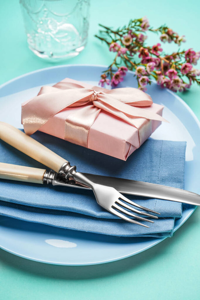 Накрытие стола с подарочной коробкой, столовыми приборами и цветочным декором на бирюзовом фоне - Фото, изображение