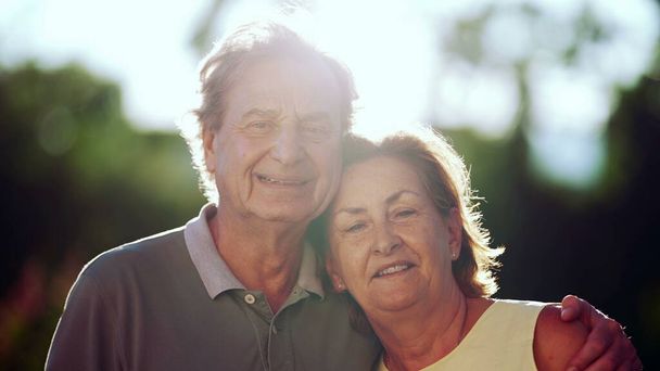 Idősebb pár mosolyog a kamera előtt a parkban. Férj arcképét, ahogy átöleli az idősebb feleséget. Háttérfény napfény - Fotó, kép