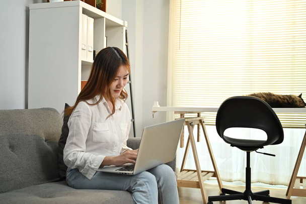 Mujer asiática joven leyendo noticias en línea, comprobando las redes sociales en el ordenador portátil en casa. Concepto de personas, tecnología y estilo de vida. - Foto, imagen