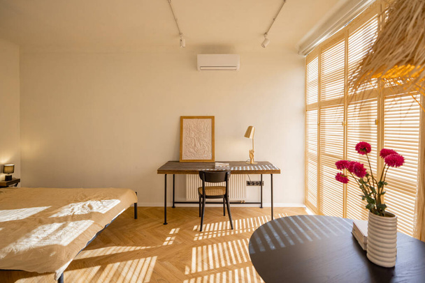Стильная и солнечная однокомнатная квартира в бежевых тонах с обеденным столом, рабочим местом и кроватью. Уютный дом и концепция дизайна интерьера. Настоящее фото - Фото, изображение