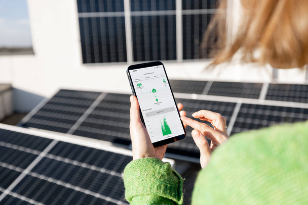 Kadın, güneş enerjisi santralinden enerji üretimini cep telefonuyla takip ediyor. Çalışan programla telefon ekranında yakın çekim görüntüsü. Güneş enerjisi üretiminin uzaktan kontrol kavramı - Fotoğraf, Görsel