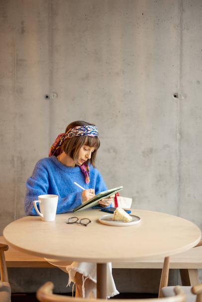 Young adult woman works on a digital tablet while sitting at modern coffee shop. Concept of remote creative work online. Concrete wall on background - Foto, Imagen