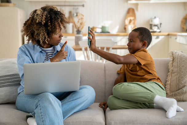 Konzentrierte schwarze freiberufliche Mutter, die zu Hause auf der Couch sitzt und am Laptop arbeitet. Kleiner afroamerikanischer Sohn lenkt von der Arbeit ab, zeigt ihr Smartphone, macht Lärm und bittet die umtriebige Mutter um Aufmerksamkeit.  - Foto, Bild