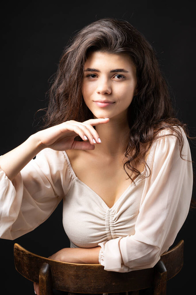 Classic studio portrait of a young brunette in a white loose top, who is sitting on a chair against a black background. - Foto, imagen
