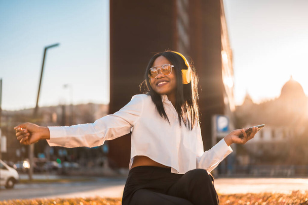 African american woman listening to music with headphones at sunset in the city, music online and dancing - Photo, Image