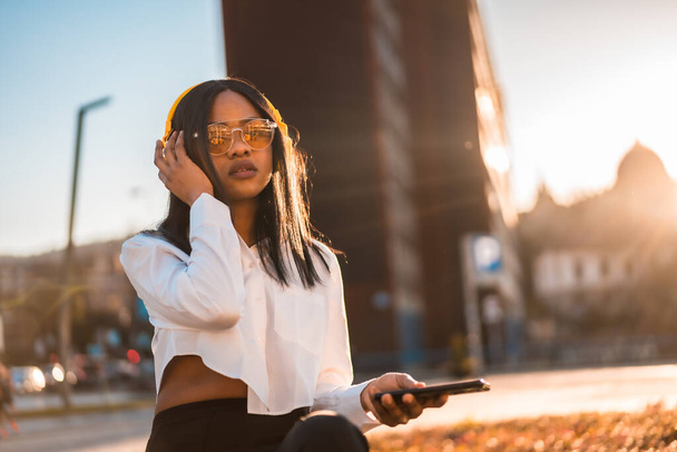 African american woman listening to music with headphones at sunset in the city, online music - Foto, Imagem