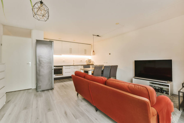 una sala de estar con paredes blancas y suelos de madera, incluyendo un sofá naranja frente al televisor - Foto, imagen