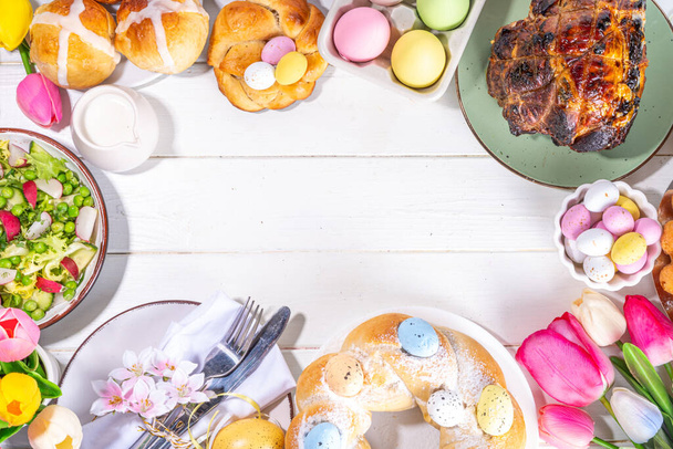 Festive served Easter dinner or brunch table, with Easter egg, tulip flower bouquet, traditional Easter foods - cake, cross buns, wreath bread, glazed ham and spring salad, white table background - Foto, Imagen
