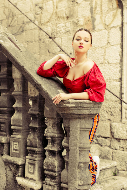 A beautiful girl in red is standing on the old stairs to the castle. Woman in accessories and professional make-up. Fashion and style - Photo, Image