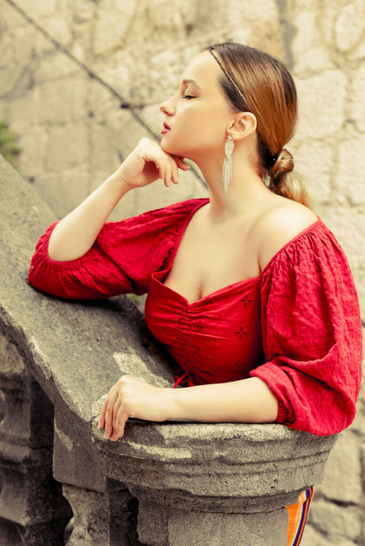 Een mooi meisje in het rood staat op de oude trap naar het kasteel. Vrouw in accessoires en professionele make-up. Mode en stijl - Foto, afbeelding