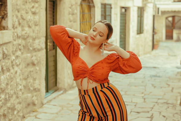 Beautiful stylish girl with long earrings, wearing an orange suit looks at the camera. Traveling around Europe Fashionable woman smiling in the old town and posing.  - Photo, image