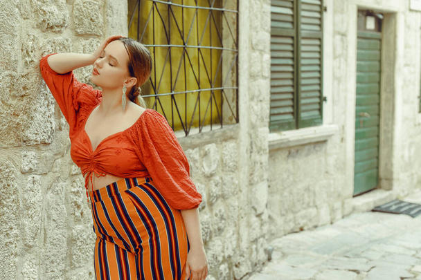 Woman fashion in the old city and posing. Beautiful stylish girl with long earrings and orange suit near the door. Traveling around Europe. - Φωτογραφία, εικόνα