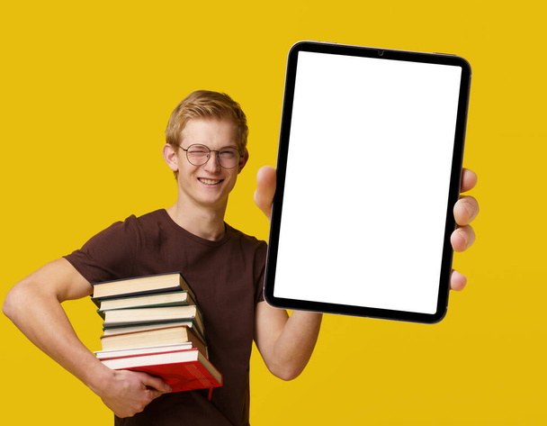 Young student holding a tablet PC with a white blank screen, surrounded by books, studying and using modern technology for academic purposes. Blond man smiles with PDA isolated on yellow background - 写真・画像