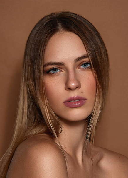 Close-up portrait of beautiful young girl with straight blonde hair, blue eyes and natural makeup posing against light brown studio background. Concept of female beauty, fashion, modern style, youth - Fotó, kép