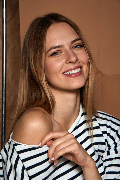 Natural beauty and femininity. Portrait of young beautiful girl in striped shirt smiling, posing against light brown background. Concept of female beauty, fashion, modern style, youth - Fotó, kép