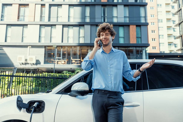 Progressive businessman talking on the phone, leaning on electric car recharging with public EV charging station, apartment condo residential building on the background as green city lifestyle. - Foto, immagini