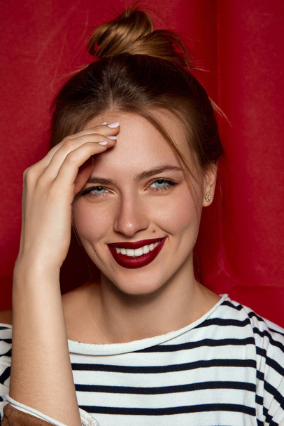 Positive femininity. Portrait of young beautiful girl with red lips and hair bun, in striped shirt posing against red studio background. Concept of female beauty, fashion, modern style, youth - Foto, Imagem