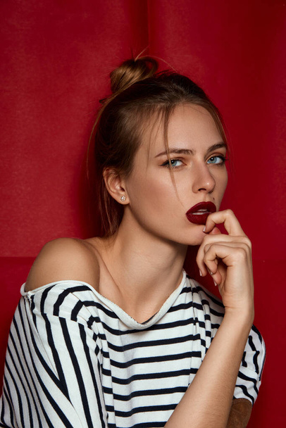 Portrait of young beautiful girl with blue eyes, red lips and hair bun, in striped shirt posing against red studio background. Concept of female beauty, fashion, modern style, youth - Foto, Bild