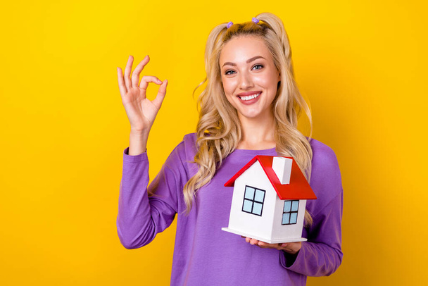 Photo of charming confident woman wear violet shirt holding small house showing okey gesture isolated yellow color background. - Фото, изображение