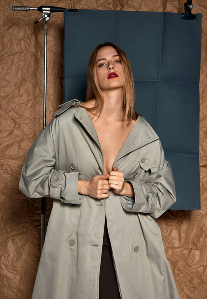 Sensuality and femininity. Portrait of beautiful young blonde girl with red lips posing in grey trench coat against studio background. Concept of female beauty, fashion, modern style, youth - Foto, Imagem