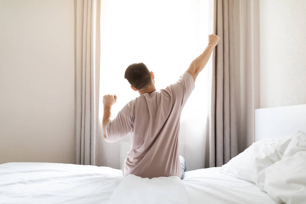 Rear view of unrecognizable man in pajamas sitting on bed and stretching body after waking up in the morning, looking at window, copy space. Comfortable healthy sleep concept - Zdjęcie, obraz