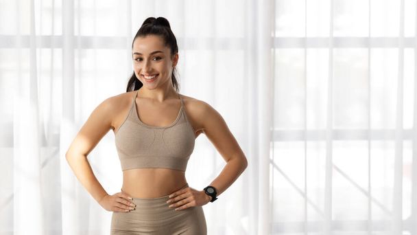 Sports lifestyle, fitness, gym concept. Portrait of sporty attractive young woman in beige sportswear with smart watch on her wrist smiling at camera, standing by window, copy space, banner - Фото, изображение