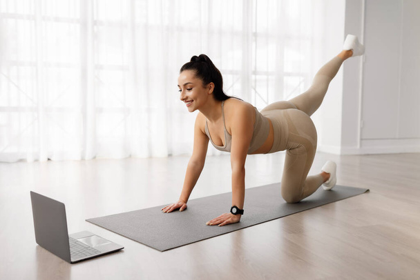 Online workout. Sporty well-fit beautiful brunette young woman in sportswear exercising on yoga mat at home, stretching legs, watching fitness video on pc laptop and smiling, copy space - Foto, Imagen
