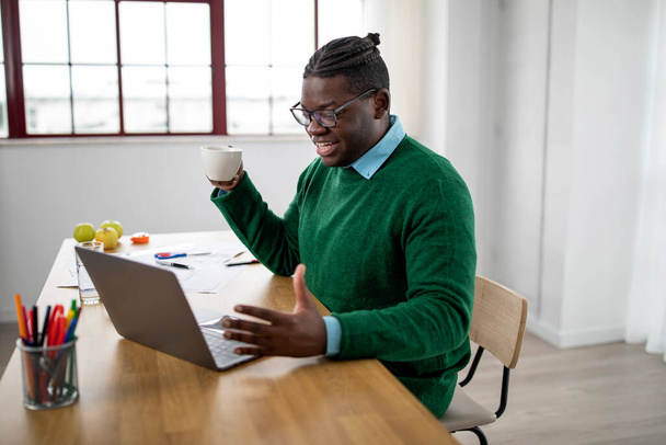 Веселый черный человек за ноутбуком Общение решений видео-звонок проведение кофе Кубок, работа в Интернете сидя за столом в современном офисе. Концепция удаленного делового общения. Шот с видом сбоку - Фото, изображение