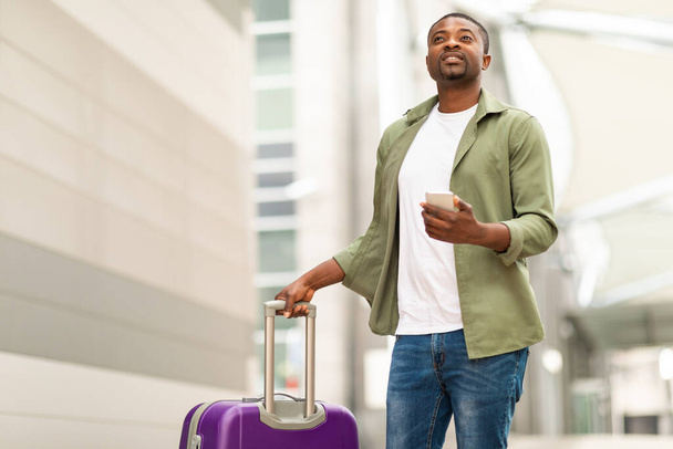 African American Man utilisant le téléphone portable et l'application de voyage Réservation de billets bon marché debout avec valise à l'aérogare moderne à l'extérieur. Tourisme et technologie Concept - Photo, image