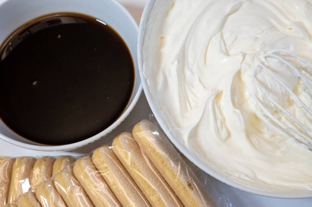 zbliżenie składników tiramisu: filiżanka kawy, opakowanie herbatników savoiardi i serek mascarpone soft cream w misce, proces gotowania w kuchni   - Zdjęcie, obraz
