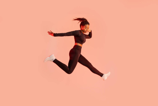 Studio shot of fit young black woman jumping on peach neon light studio background, free space. African american lady in sportswear exercising and jumping in air - Photo, Image