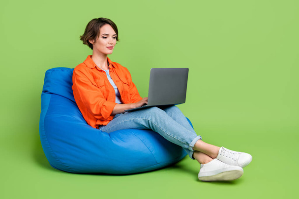Full length photo of cheerful woman wear denim jacket bean bag typing emails gadget empty space isolated green color background. - Foto, immagini