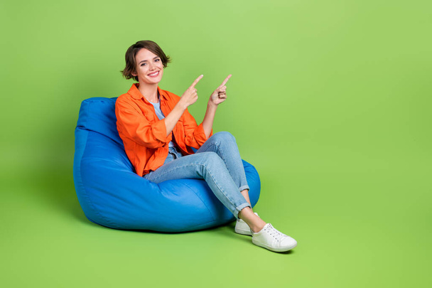 Full length photo of adorable woman wear denim jacket bean bag pointing two fingers empty space isolated green color background. - Zdjęcie, obraz