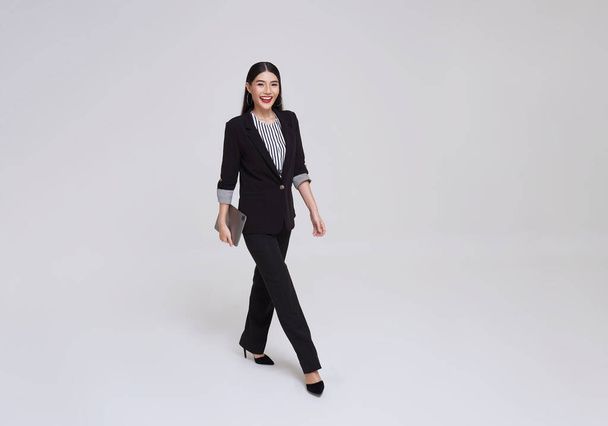 幸せなアジアのビジネス女性笑顔で正式なスーツ保持タブレットと上を歩くグレーの背景. - 写真・画像