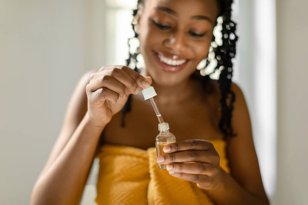 Black lady holding open bottle with moisturizing face serum, sitting in bedroom and smiling, enjoying making daily skin care treatments, selective focus, closeup - Фото, зображення