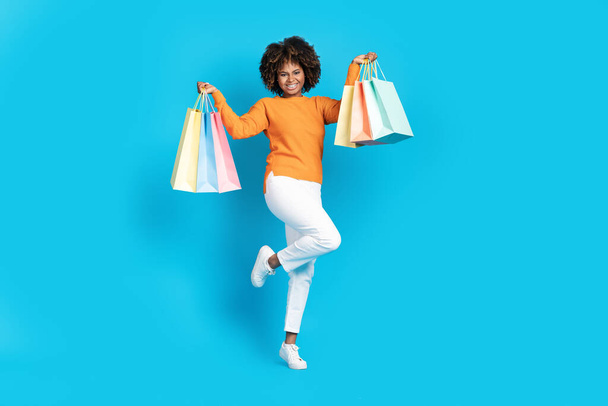 Cheerful happy stylish young black woman with bushy hair shopaholic dancing with colorful shopping bags in her hands over blue studio background, celebrating black friday deals, copy space - 写真・画像