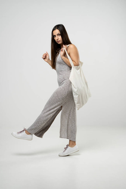 Brunette woman in grey dress holding cotton shopper bag with vegetables, products in white room. Eco friendly shopping bags. Zero waste, plastic free concept - 写真・画像