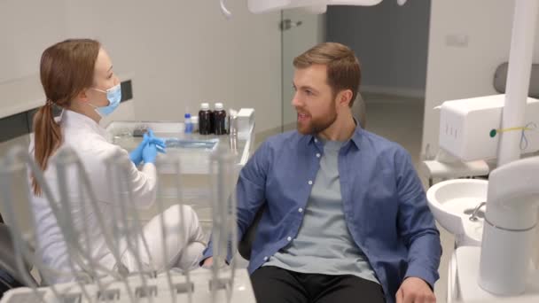 Blonde female dentist in dental office talking with male patient and preparing for treatment. Handsome bearded man in dentist chair looking at his doctor with smile - Imágenes, Vídeo